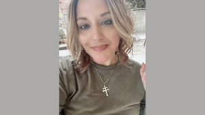 Female with highlighted hair and a relaxed expression, photographed outdoors with a fence and greenery in the background.