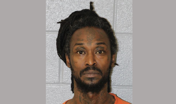 Image of a man with a full beard, tattoos, and dreadlocks, shown from the shoulders up wearing an orange outfit, photographed against a light gray background.