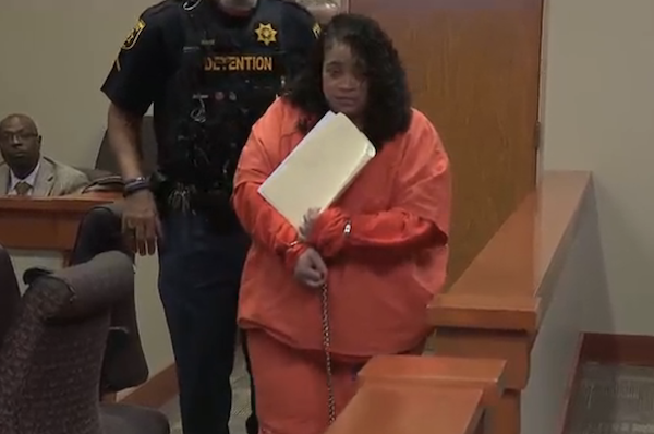 In a courtroom setting, a woman dressed in an orange jumpsuit and restrained at the wrists appears before a judge, accompanied by a detention officer.