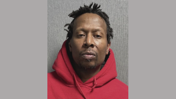 Close-up image of a man facing forward, wearing a red hoodie, with a neutral facial expression and a gray wall behind him.