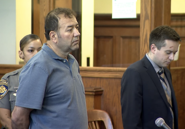 Three individuals stand at the front of a courtroom; the foreground man appears to be the defendant, while a uniformed officer and a man in a suit stand nearby.