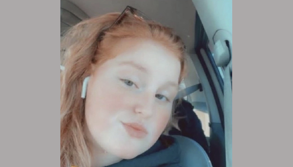 Portrait of a young person with soft facial features and wavy hair, wearing a black jacket and AirPods, photographed from inside a vehicle.