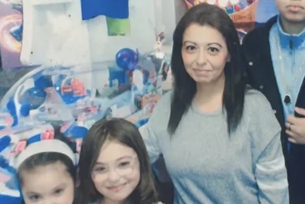 An indoor scene features a woman standing between two children, in front of a wall covered with toys and artwork, the child on the left smiling broadly and the child on the right wearing a necklace.
