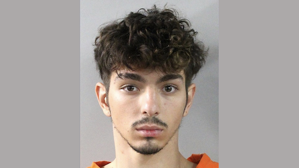 Portrait of a young adult male with curly hair, light facial hair, and an orange shirt, photographed in front of a simple, light-colored backdrop.