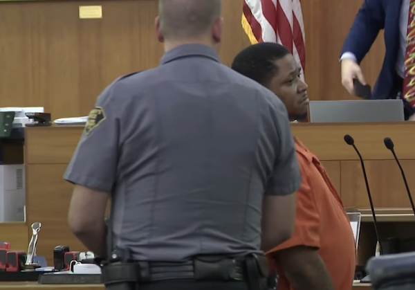 A courtroom image featuring a person in orange jail clothing, a uniformed guard, and another official near the judge’s bench, with part of the U.S. flag visible in the background.