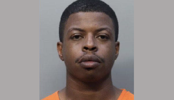 A solemn-looking man in an orange garment stares directly forward, centered against a neutral-colored wall.