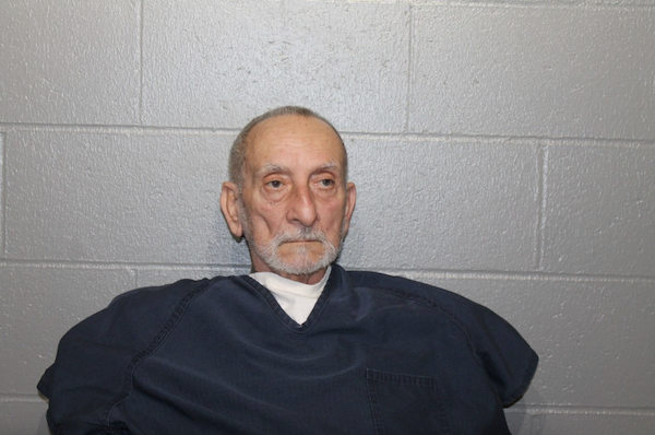 Portrait of an older individual dressed in jail attire, seated and unsmiling, with a gray brick wall as the backdrop.