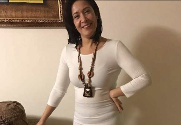 Smiling woman with shoulder-length dark hair wearing a white dress and beaded necklace poses in a living room, with her left hand on her hip and a painting on the wall behind her.
