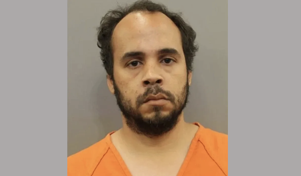 Man wearing an orange prison jumpsuit shown against a plain background, looking directly at the camera with a neutral expression.