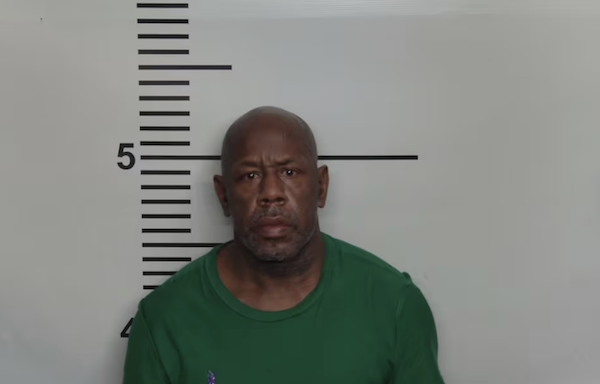 A man with a bald head and trimmed beard is photographed against a wall featuring a height ruler, dressed in a green crewneck top.