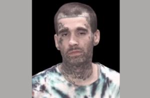 Close-up of a person with dark hair, visible facial and neck tattoos, and a tie-dye shirt, centered on a black background.