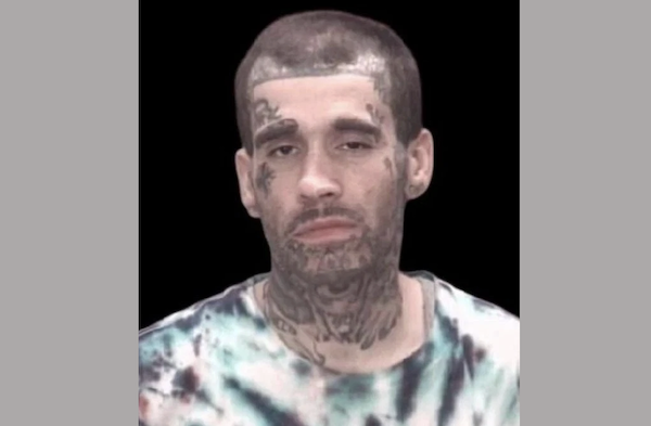 Close-up of a person with dark hair, visible facial and neck tattoos, and a tie-dye shirt, centered on a black background.