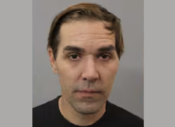 Head-and-shoulders photo of a man with a side-part hairstyle, slight stubble, and a somber expression, photographed against a neutral gray background.