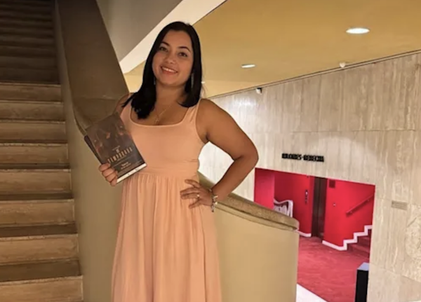 Woman wearing a pastel dress poses inside a brightly lit building, holding a book and standing near a curved stairway railing.