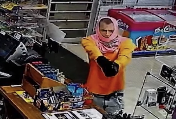 Store surveillance photo depicting an individual with a makeshift head covering and heavy clothing, extending their arms toward the camera in what looks like a confrontation.