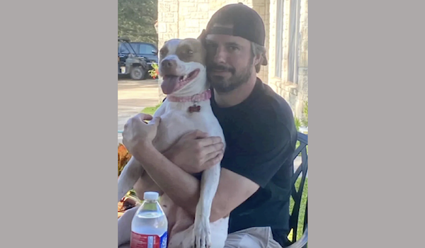 Outdoors, a cheerful dog with its mouth open is hugged by a man dressed in black, the pair sitting together by what appears to be a house or building.