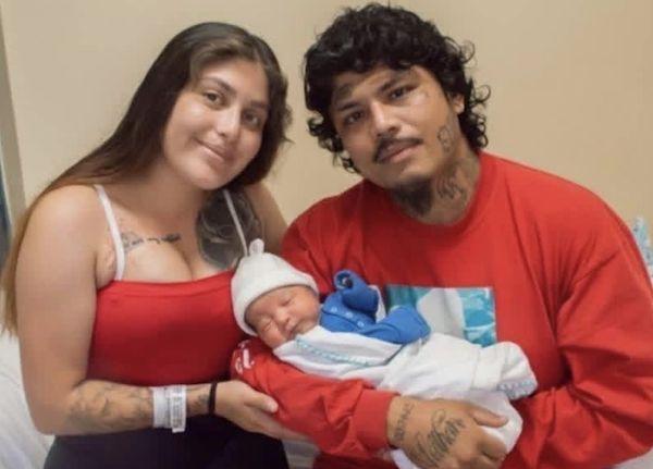 Three people are featured in the image: a woman and man, both with dark hair and tattoos, sitting closely while lovingly holding a newborn baby dressed warmly.