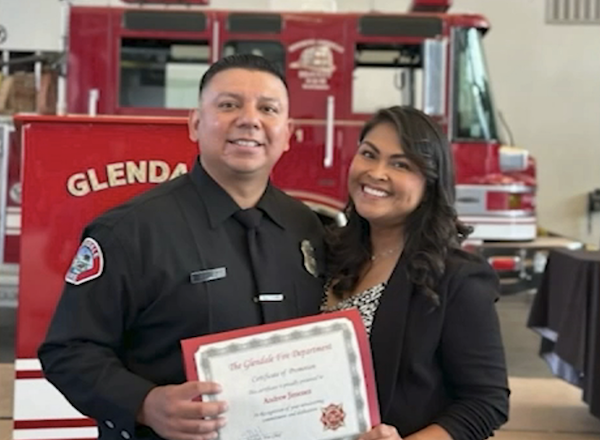 Two people smile for a photograph, with the firefighter displaying a certificate and a fire department vehicle visible behind them.