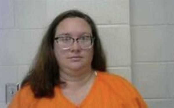 Person with straight hair, glasses, and an orange jail shirt faces the camera in a booking photo, neutral expression visible.