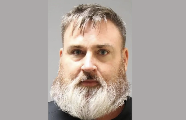 Close-up photo of a man with blue eyes, short brown-gray hair, and a full bushy beard, centered against a plain gray background.