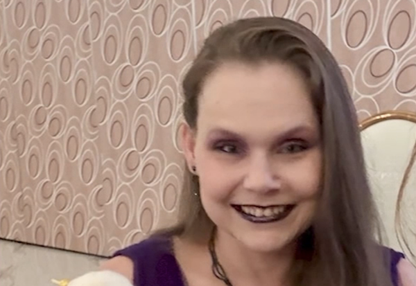 Close-up of a woman with long straight hair and bold makeup smiling in a room with beige circular-pattern wallpaper.