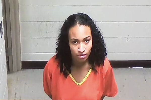 Booking photo shows a woman in an orange jail uniform with hands behind her back in a holding area.
