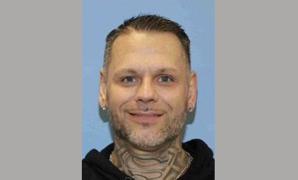 Portrait of a man with stubble, earrings, and a prominent tattoo across his neck, photographed against a solid blue background.