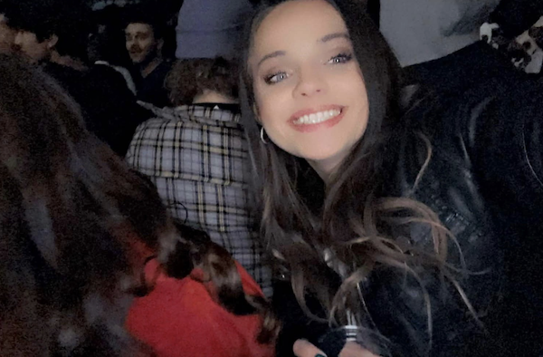 Woman grinning at the camera while taking a selfie in a crowded bar or party environment with dim lighting and people behind her.