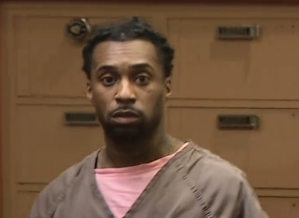 Indoor photo of a man in custody clothing speaking or reacting, with beige walls, a calendar in the background, and an officer partly visible nearby.
