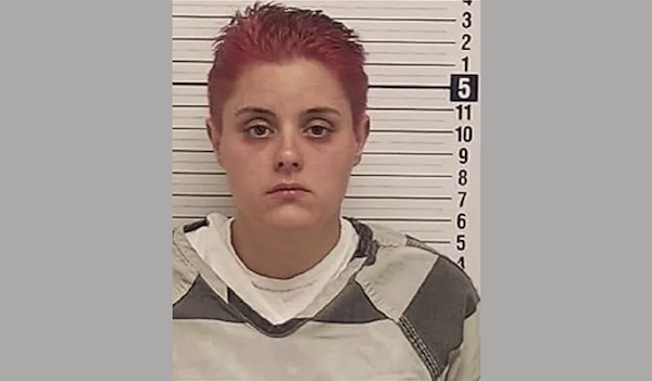 Booking image of a short-haired woman with red dyed hair, centered in frame, posed in front of measurement lines.