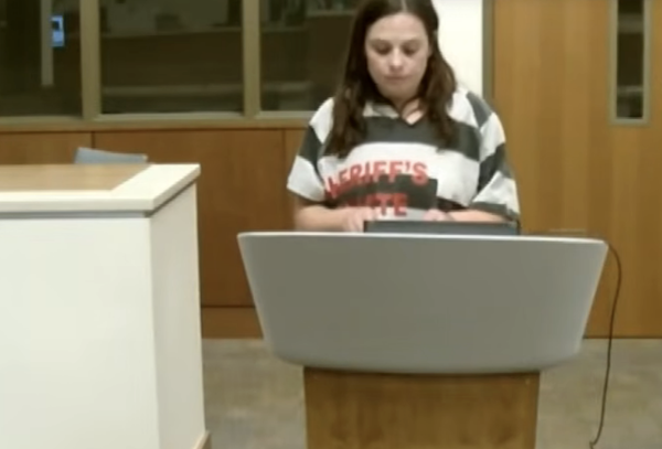 Front-facing courtroom image of a woman in a gray-and-white striped inmate uniform behind a stand.