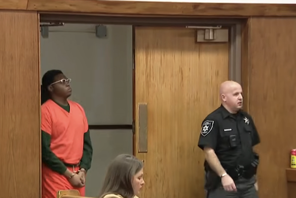 Indoor courtroom image of a man in custody wearing an orange outfit over a green shirt, flanked by officers in black uniforms.