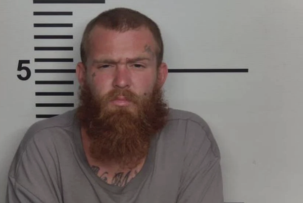 Cropped mugshot-style image showing a man in a gray shirt seated before a wall with height markings.