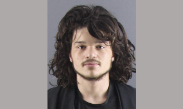 Head-and-shoulders mugshot-style image of a man with wavy hair wearing a dark shirt.