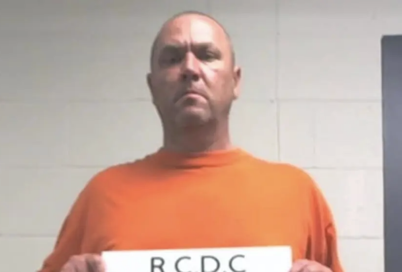 Low-resolution jail photo of a man in an orange shirt standing against a plain wall.
