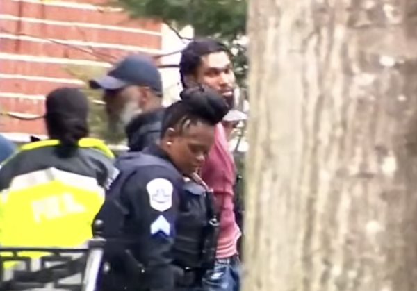 A uniformed officer walks ahead as police move a man through a narrow outdoor area.