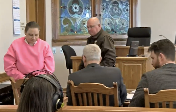 Court hearing image showing a woman in a pink top, a uniformed officer, seated legal staff and a judge’s bench in the background.