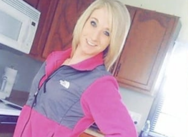 Smiling blonde woman stands with one hand on her hip in a kitchen with wood cabinets.
