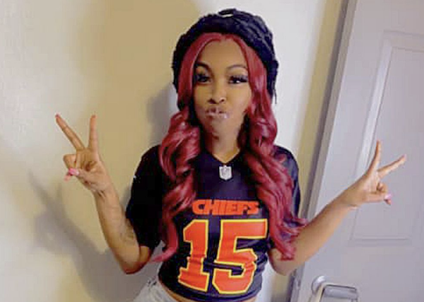 A smiling woman in Kansas City Chiefs apparel stands near a doorway and flashes peace signs.