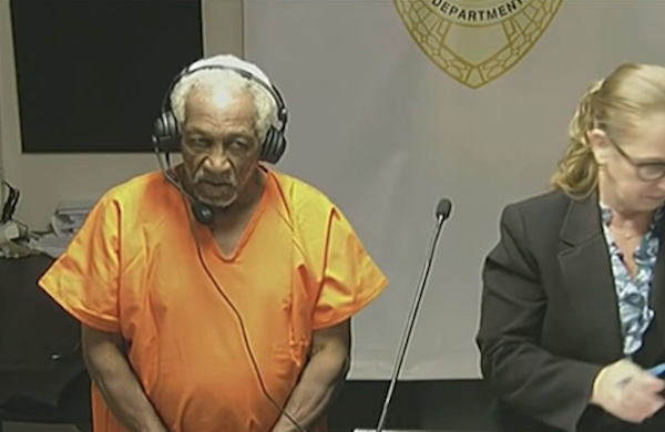 An elderly defendant in orange clothing listens through headphones during a court proceeding.