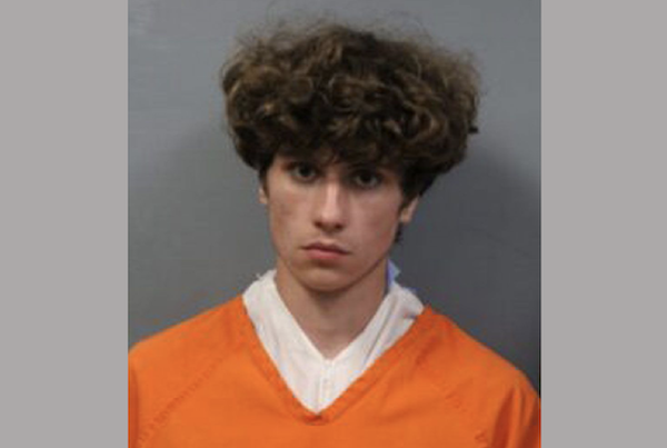 Close-up mugshot of a curly-haired young man wearing orange clothing and facing forward against a plain gray backdrop.