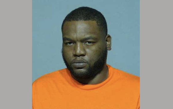 Close-up portrait of a man with short dark hair and a trimmed beard, photographed against a blue-gray backdrop in an orange shirt.