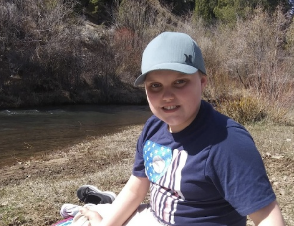 Child seated beside a creek in a natural setting, wearing a light blue cap and dark shirt with shrubs and dry vegetation behind them.
